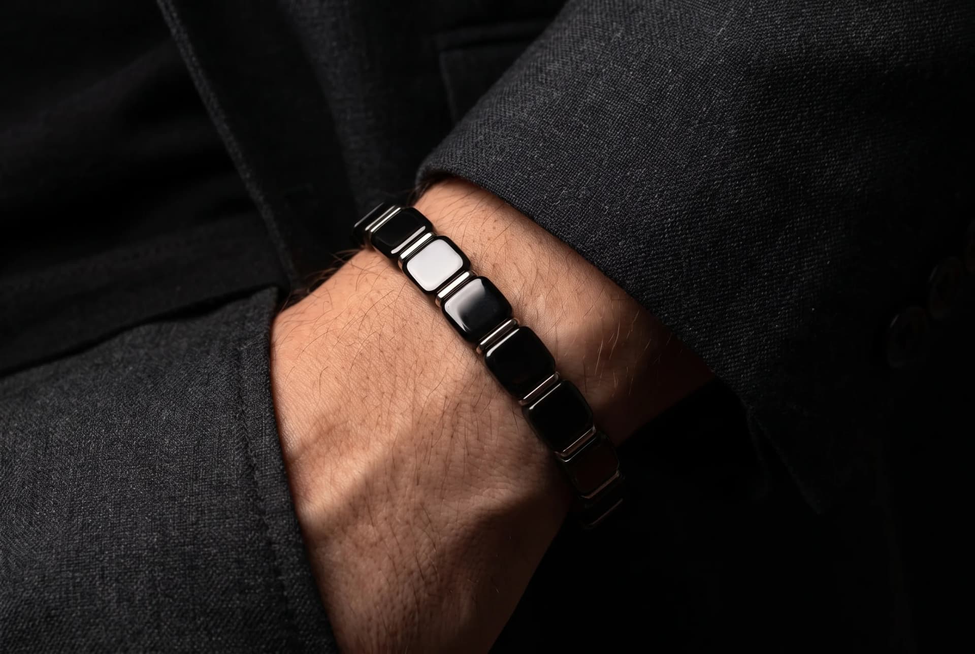 Editorial photograph of a man's wrist wearing a metallic hematite beaded bracelet with a dark suit cuff