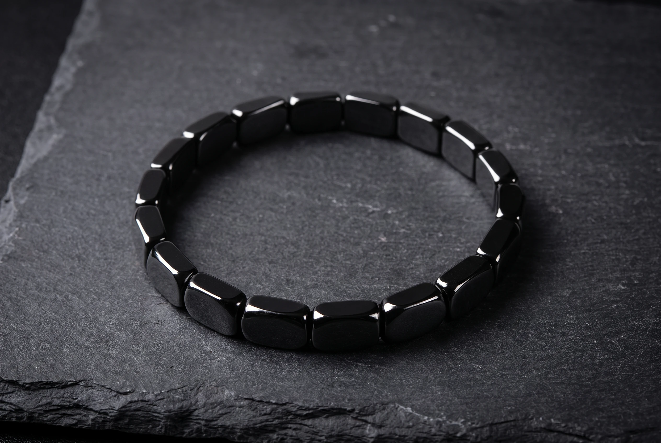 Overhead photograph of the Hematite Men's Bracelet coiled in a loose oval on a dark slate surface under soft single-source light